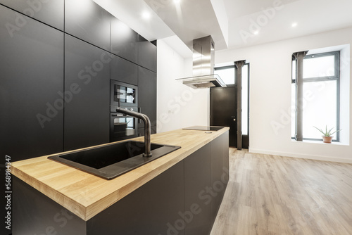 Newly installed open plan kitchen with black cabinets, appliances and faucets and an island with a wood countertop on laminate flooring, black metal door and bay windows © Toyakisfoto.photos
