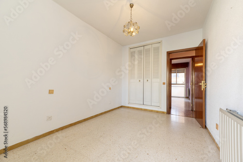 Empty room with white walls with a built-in wardrobe with white folding doors in the Venetian style, a lamp hanging from the ceiling and a smooth door made of varnished sapele wood