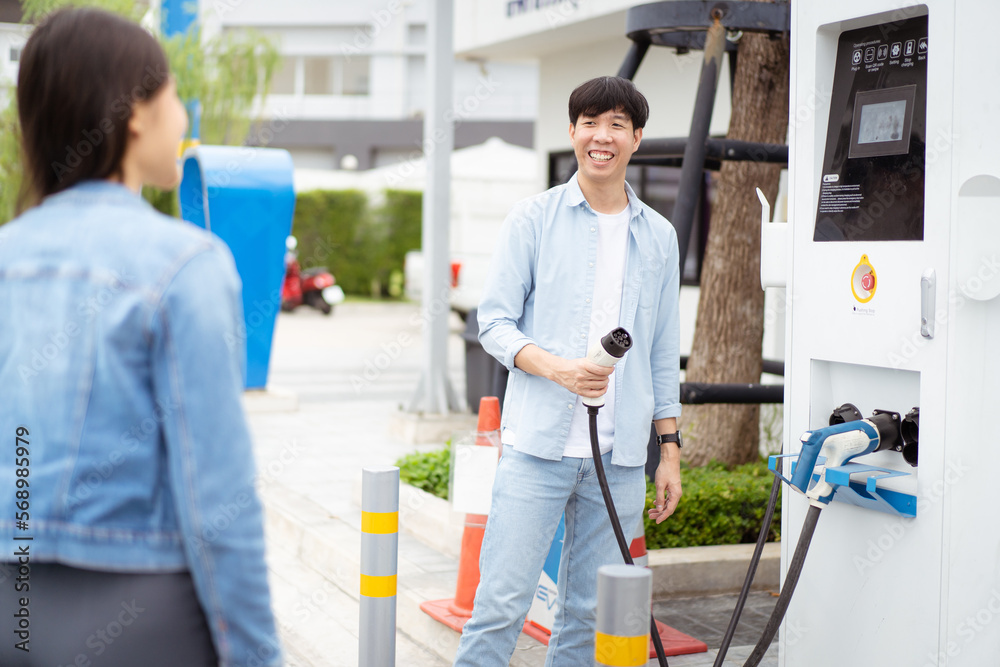 Fototapeta premium Asian man and woman holding AC type 2 EV charging connector at EV charging station. Road vehicle traveling or transportation with the clean energy concept.
