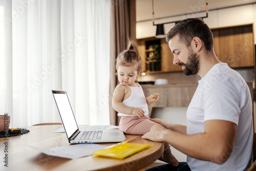A stay at home dad is working on a laptop while his toddler is helping him. photo