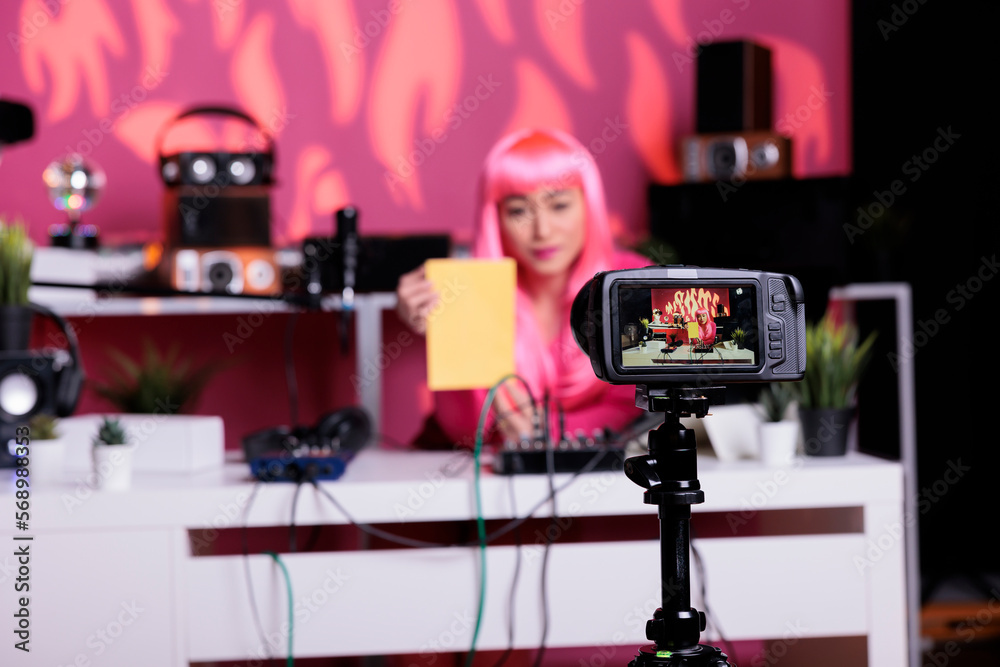 Smiling vlogger holding notebook showing product in front of camera while recording review using professional vlogging equipment. Content creator standing at desk filming podcast in broadcast studio Smiling vlogger holding notebook showing product in front of camera while recording review using professional vlogging equipment. Content creator standing at desk filming podcast in broadcast studio
