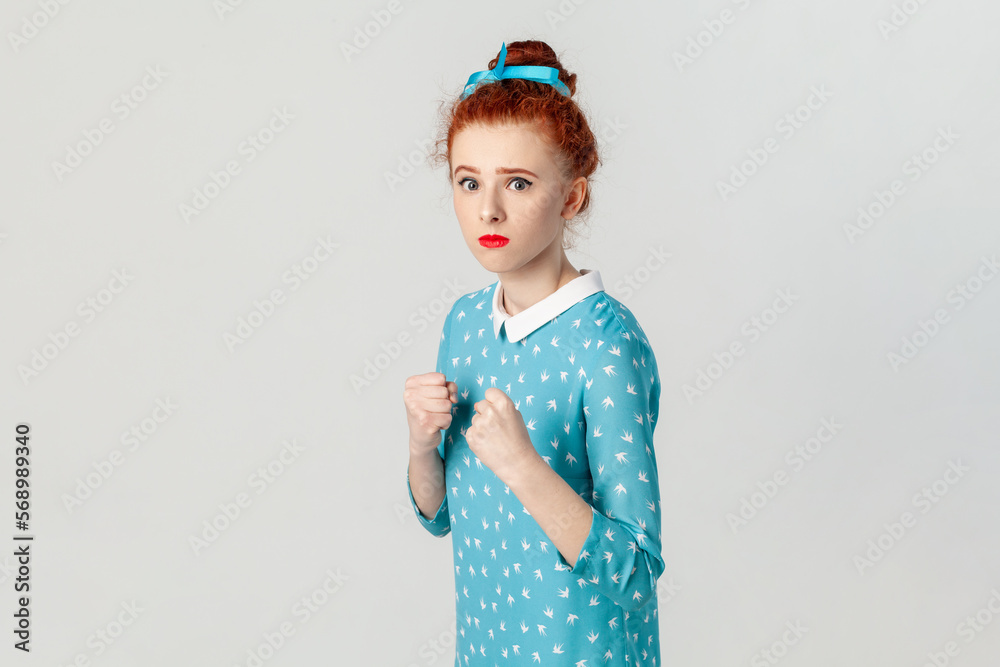 Portrait of confident serious red haired woman with hair bun, standing with clenched fists, being ready to attack, boxing, wearing blue dress. Indoor studio shot isolated on gray background. Portrait of confident serious red haired woman with hair bun, standing with clenched fists, being ready to attack, boxing, wearing blue dress. Indoor studio shot isolated on gray background.
