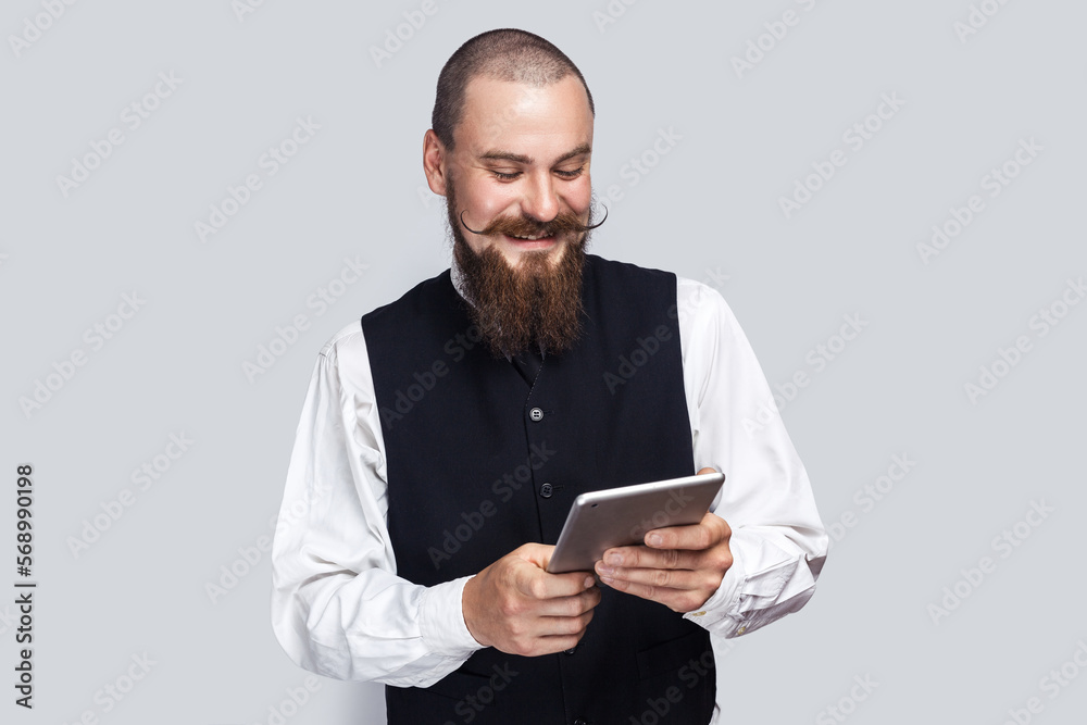 Portrait of extremely happy positive man with beard and mustache using tablet and laughing, reading jokes or watching funny video. Indoor studio shot isolated on gray background. Portrait of extremely happy positive man with beard and mustache using tablet and laughing, reading jokes or watching funny video. Indoor studio shot isolated on gray background.