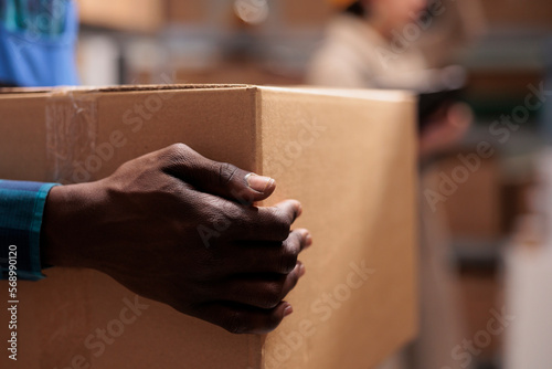 Shipping service warehouse african american man worker holding received parcel in hands. Delivery company employee lifting heavy cardboard box in storehouse with close view on arm