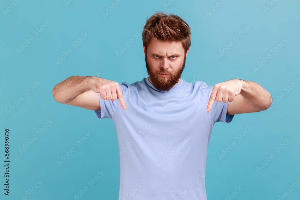 Here and right now. Portrait of bearded man standing and looking at camera with fingers down, commanding to act here and now, demanding immediate result. Indoor studio shot isolated on blue background Here and right now. Portrait of bearded man standing and looking at camera with fingers down, commanding to act here and now, demanding immediate result. Indoor studio shot isolated on blue background
