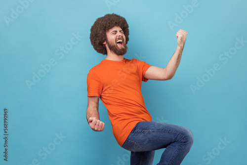 Portrait of man with Afro hairstyle in T-shirt stands with excited expression, raising fists, screaming, shouting yeah, celebrating his victory, success. Indoor studio shot isolated on blue background photo