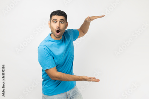 Foto Portrait of shocked astonished surprised unshaven man wearing blue T- shirt standing showing advertisement area between his arms, copy space, big size