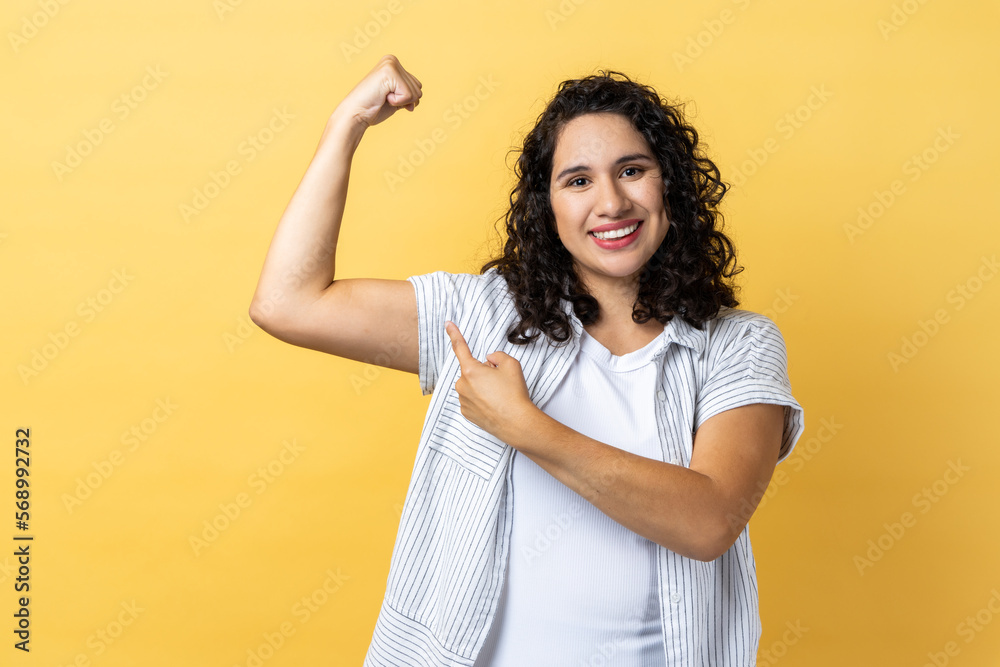 Portrait of woman with dark wavy hair showing her arm muscle and point it with finger, proud of her strength and leadership skills. Indoor studio shot isolated on yellow background. Portrait of woman with dark wavy hair showing her arm muscle and point it with finger, proud of her strength and leadership skills. Indoor studio shot isolated on yellow background.