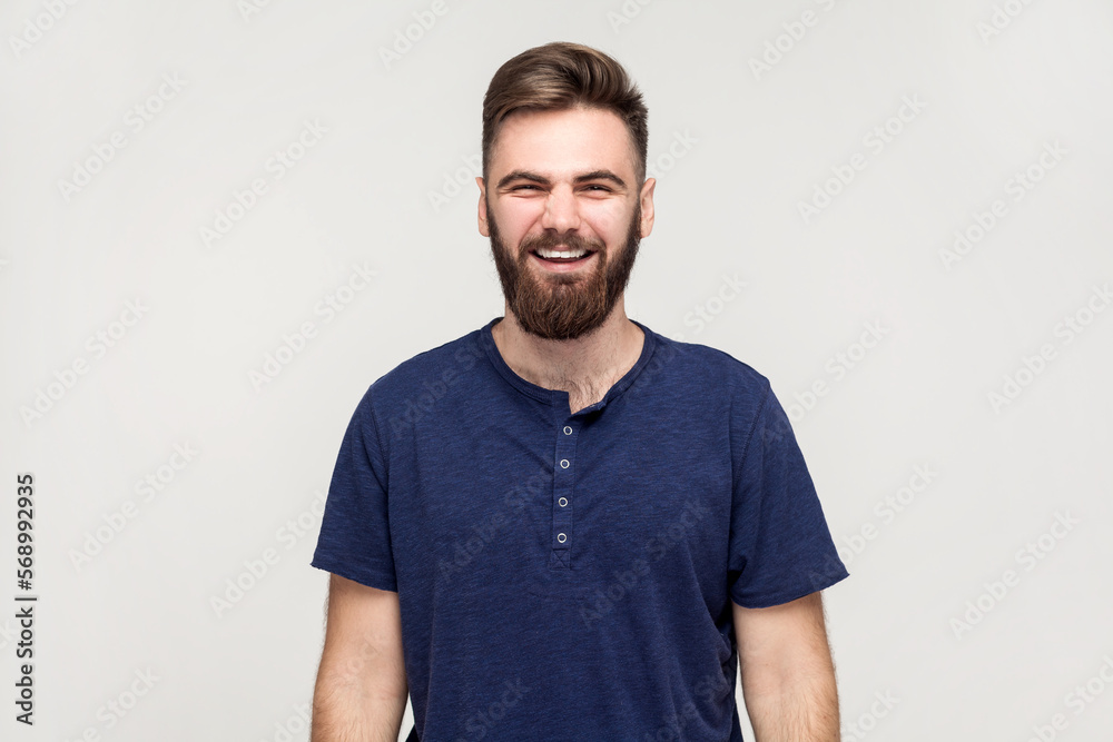 Obraz premium Portrait of overjoyed extremely happy handsome man with beard wearing dark blue T-shirt hunching from laughter, laughing out loud, crazy face. Indoor shot isolated on gray background.