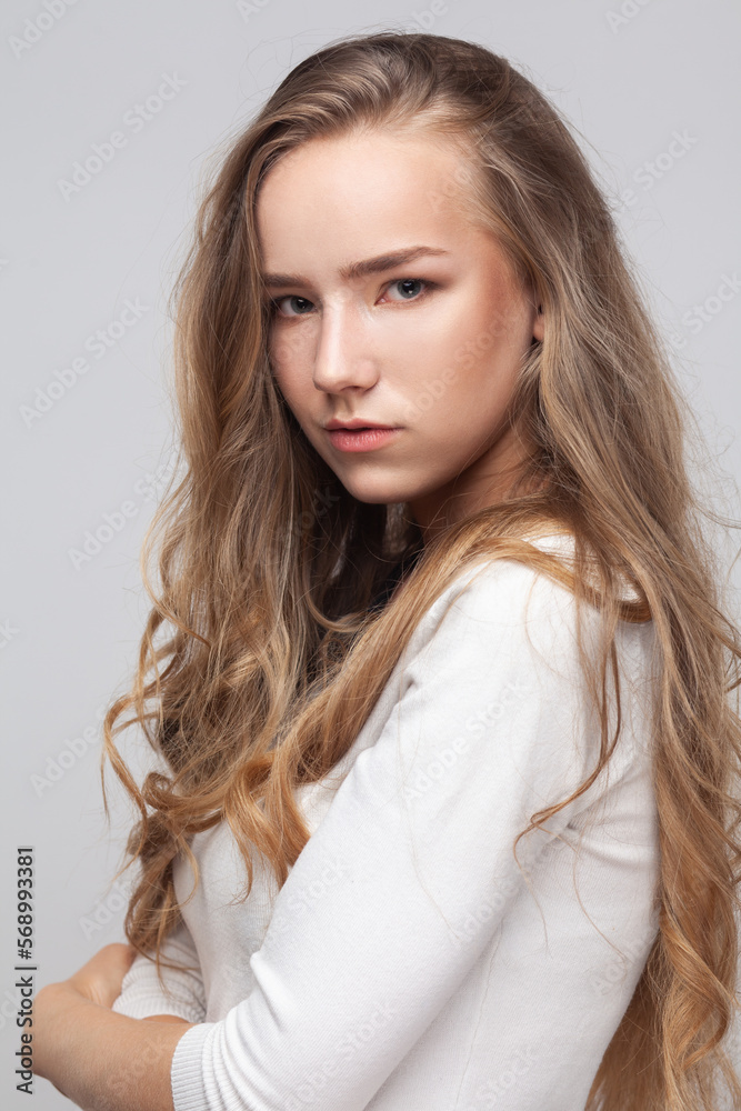 Closeup portrait of beautiful attractive young adult woman with long curly blond hair looking at camera with pensive facial expression. Indoor studio shot isolated on gray background. Closeup portrait of beautiful attractive young adult woman with long curly blond hair looking at camera with pensive facial expression. Indoor studio shot isolated on gray background.