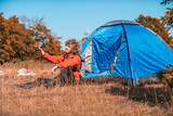 A man with a tent enjoys a sunny day whileusing a smartphone and camping in the mountains