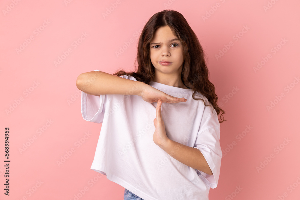 Portrait of serious concentrated little girl wearing white T-shirt showing time out gesture, looking with strict expression, deadline. Indoor studio shot isolated on pink background. Portrait of serious concentrated little girl wearing white T-shirt showing time out gesture, looking with strict expression, deadline. Indoor studio shot isolated on pink background.
