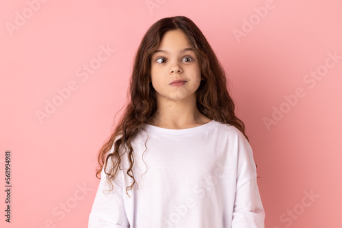 Portrait of little girl wearing white T-shirt crossing her eyes looking crazy and stupid, fooling around, having fun, vision problems. Indoor studio shot isolated on pink background. photo