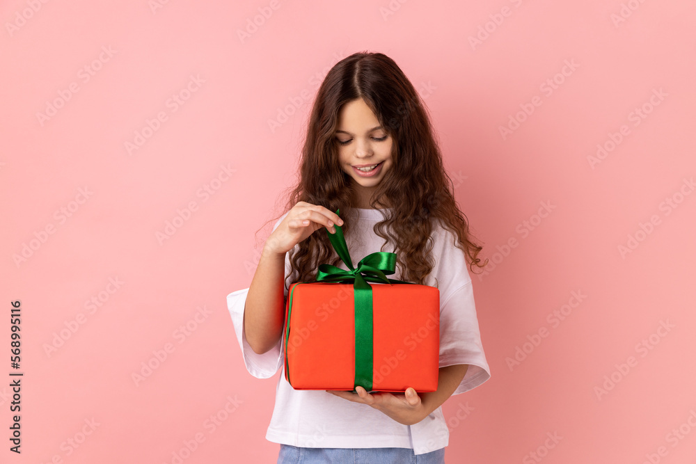 Portrait of little girl wearing white T-shirt opening gift box, unwrapping birthday present for celebrating holiday, pulling ribbon, looking at gift. Indoor studio shot isolated on pink background. Portrait of little girl wearing white T-shirt opening gift box, unwrapping birthday present for celebrating holiday, pulling ribbon, looking at gift. Indoor studio shot isolated on pink background.