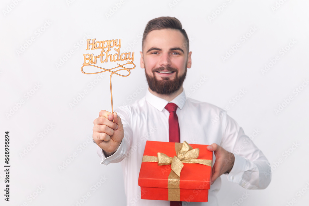 Portrait of smiling satisfied handsome man in white shirt holding party props with happy birthday inscription and red present box. Indoor studio shot isolated on gray background. Portrait of smiling satisfied handsome man in white shirt holding party props with happy birthday inscription and red present box. Indoor studio shot isolated on gray background.