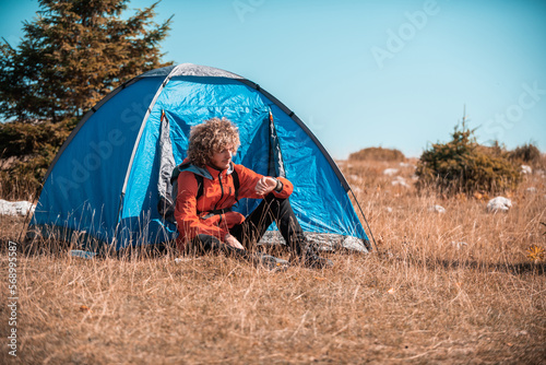 A man camping with a tent. He is surrounded by a beautiful mountain landscape, and the sun shines on his face, foreshadowing a beautiful and sunny day. The man looks satisfied and fulfilled
