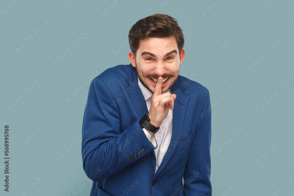 Portrait of funny smiling handsome man with mustache standing with finger near lips showing shh gesture, keeps secret wearing official style suit. Indoor studio shot isolated on light blue background.