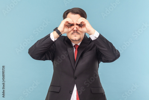 Portrait of serious concentrated man with mustache looking at camera through binocular making with hands, wearing black suit with red tie. Indoor studio shot isolated on light blue background.