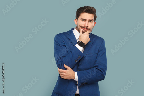 Pensive thoughtful handsome man standing and holding his chin, thinking about future, looking away, wearing white shirt and jacket. Indoor studio shot isolated on light blue background.