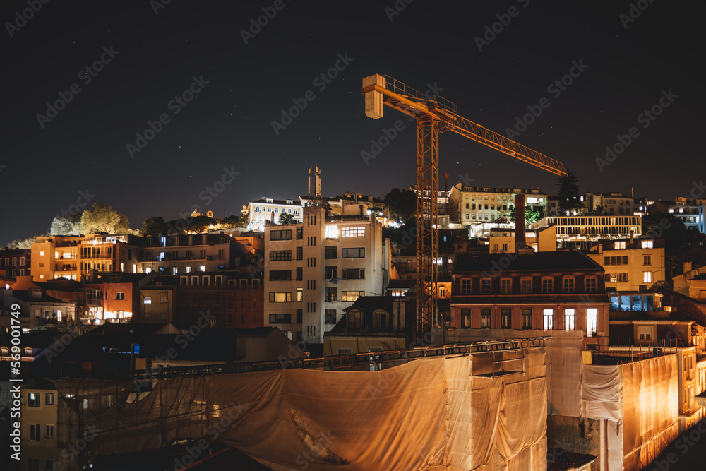 A top-down view of a city under construction at night in low-key and long exposure. Old residential houses surround the on-site building, a tall yellow crane towering over the buildings standing out