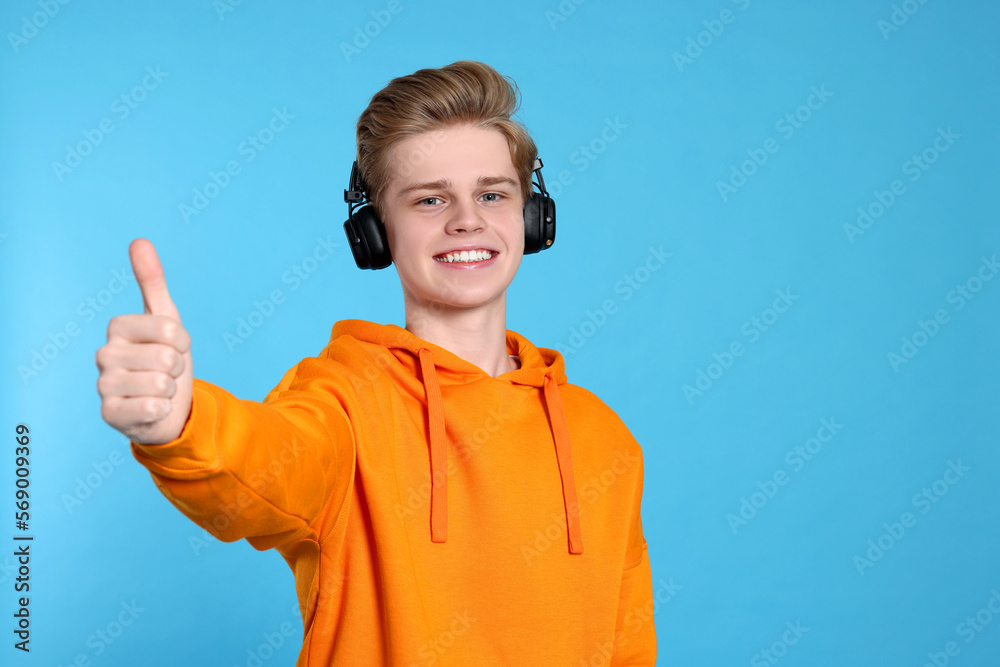 Teenage boy with headphones showing thumb up on light blue background. Space for text Teenage boy with headphones showing thumb up on light blue background. Space for text