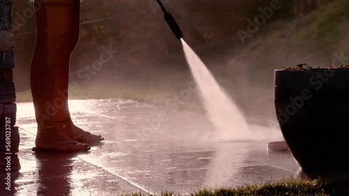 SLOW MOTION, CLOSE UP: Woman washes terrace floor with high pressure cleaner. Sunny day for outdoor spring cleaning of backyard surfaces with pressure washer. Use of powerful water jet in golden light photo