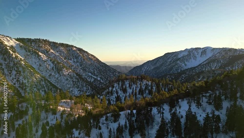 Aerial shot over Big Bear, California with snowy mountains and green pine trees during sunset.