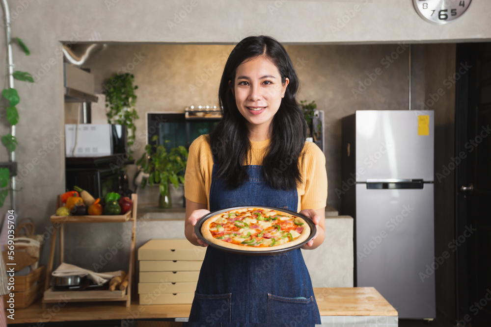 Portrait of young Attractive Asian woman in apron holding homemade fresh pizza with satisfied smiley face. Home kitchen small business food delivery.