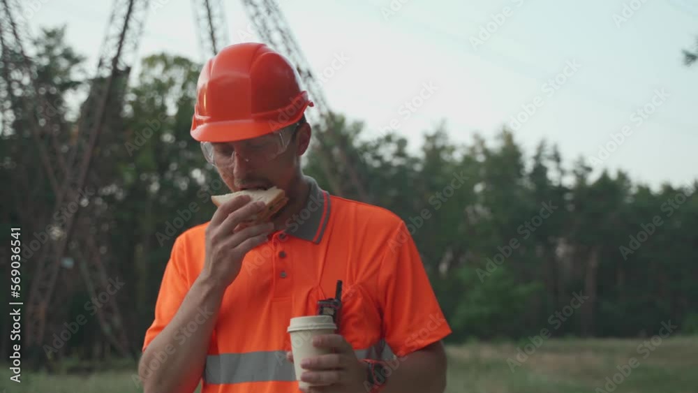 Hungry worker in overalls in electric power business eats sandwich and drinks takeaway coffee at workplace near power lines. Power worker eating snack with hot drink near high voltage power line. Hungry worker in overalls in electric power business eats sandwich and drinks takeaway coffee at workplace near power lines. Power worker eating snack with hot drink near high voltage power line.