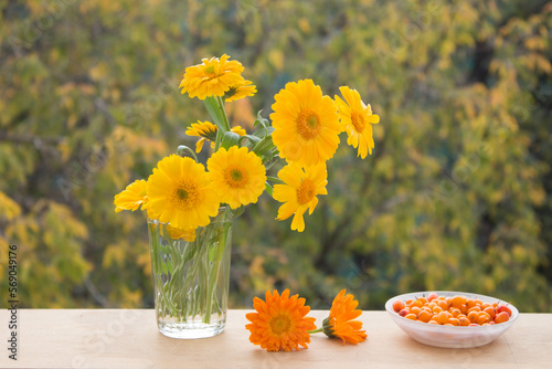 Fresh yellow flowers in glass vase and berries of sea ​​​​buckthorn in saucer. Autumn colorfull сomposition. Outdoors.