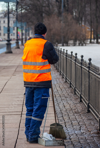 Migrant janitor in bright vest rests while working on the sidewalk.
