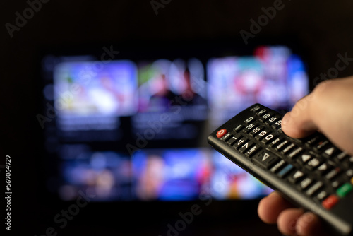 A person holds a remote control in his hand and watches TV with a blurry TV screen in the background. Viewing news