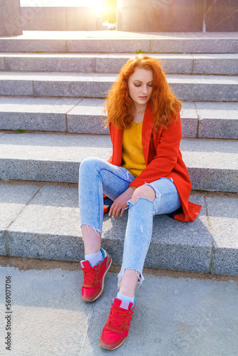 Young adult redhead woman in red coat, blue jeans and red sneakers sits on the steps of a concrete staircase. Daylight, everyday lifestyle