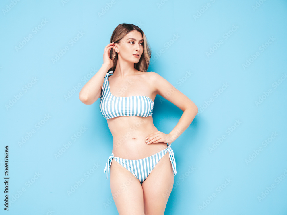 Fototapeta premium Young blond model in summer striped swimwear bathing suit. Sexy carefree woman having fun and going crazy. Female posing near blue wall in studio. Cheerful and happy. Perfect body. Slim, fit