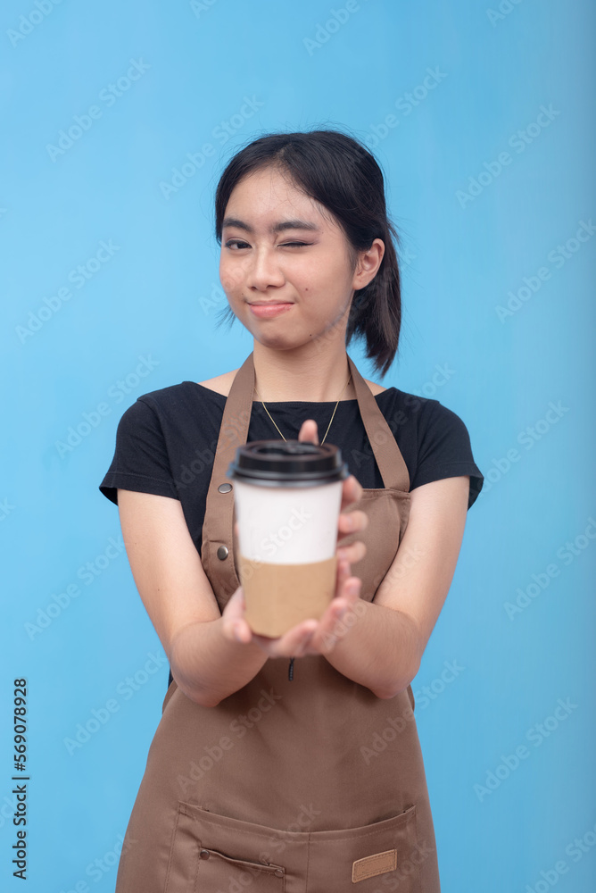 A young and cute asian female barista winks while serving a cup of coffee. Wearing a brown apron and black shirt welcoming a customer and against a light blue background. A young and cute asian female barista winks while serving a cup of coffee. Wearing a brown apron and black shirt welcoming a customer and against a light blue background.