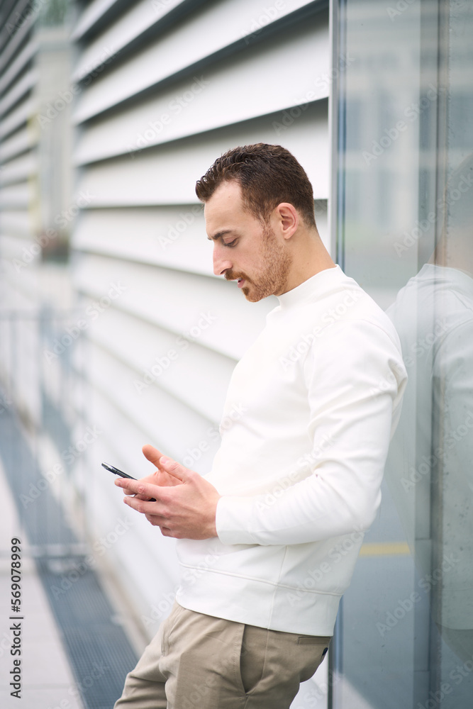 Fototapeta premium Vertical view of the serious businessman checking email, watching videos near building wall. Young stylish man chatting, reading text message on the street. Shopping online concept 
