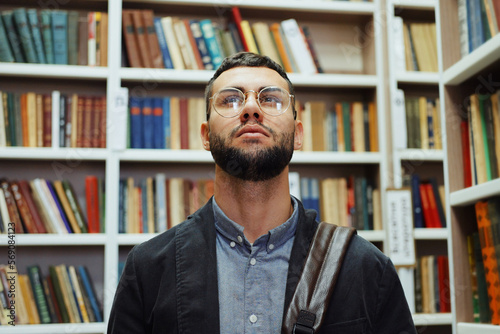 Bearded young man with eyeglasses wearing shirt and jacket standing among bookcases and looking up. Student choosing book in library. Concept of education