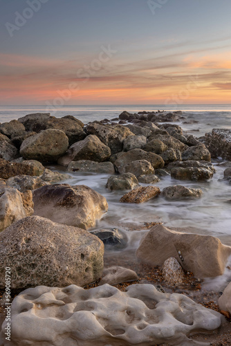 Ocean scene with low sun on the horizon, some rocks in foreground and mid ground with blurred waves around them plus a few colourful clouds.