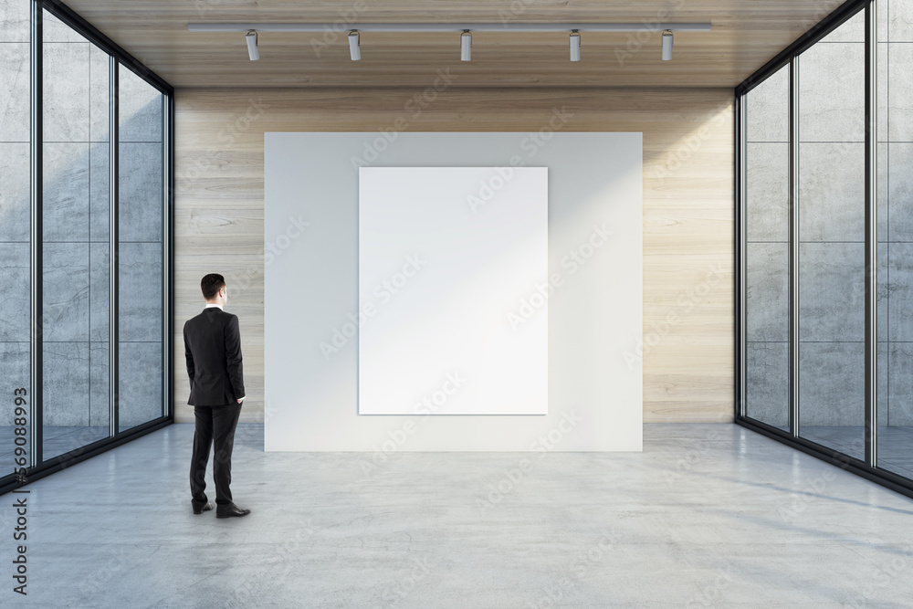 Man in black suit back view looking at blank white poster with space for your logo or text on light partition on light wooden background in empty exhibition hall with transparent walls, mock up Man in black suit back view looking at blank white poster with space for your logo or text on light partition on light wooden background in empty exhibition hall with transparent walls, mock up