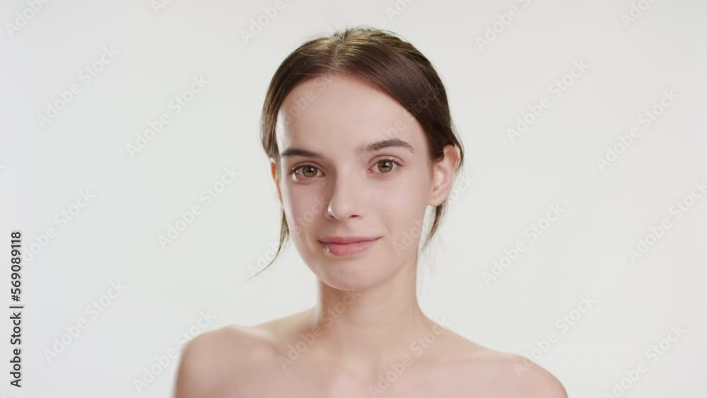 Cute young slim Caucasian woman with long brown hair strokes her face using cotton pad looking at the camera and smiling for it on grey background | Pure facial skin concept Cute young slim Caucasian woman with long brown hair strokes her face using cotton pad looking at the camera and smiling for it on grey background | Pure facial skin concept