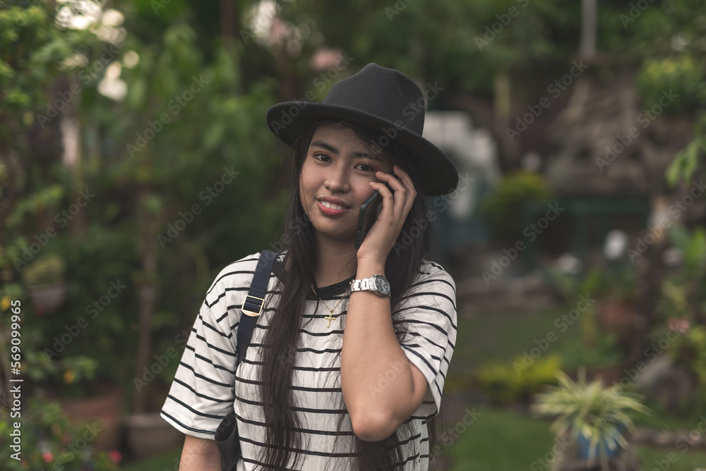 A pretty and stylish young asian woman with long straight hair talking to a friend over the phone. Wearing a white striped blouse, plain shorts and a black hat at the garden.