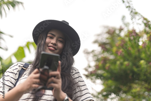 A pretty and stylish young asian woman with long straight hair and a black fedora hat looking at the camera while chatting on the cellphone. Outdoor scene.