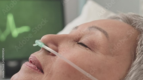Closeup of mature Asian woman with nasal cannula lying in hospital bed, rehabilitating after surgery operation photo