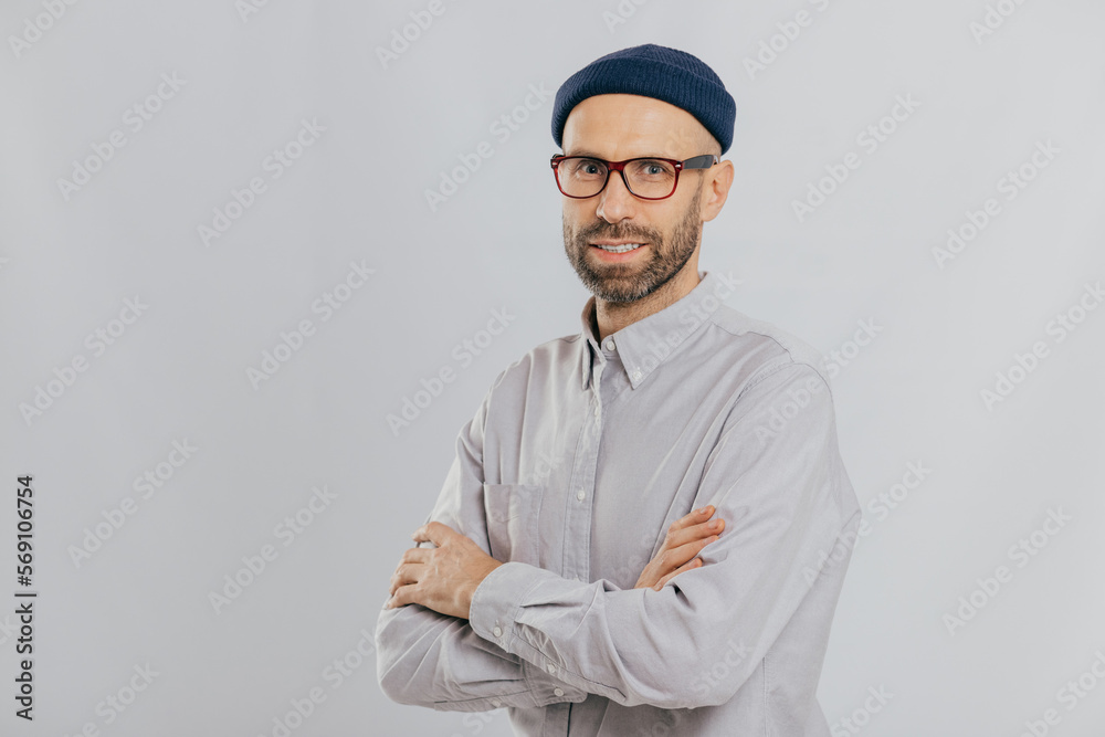 Isolated shot of pleasant looking unshaven male keeps hands crossed, wears stylish hat, optical glasses, models over white background with copy space for your advertising content, enjoys leisure Isolated shot of pleasant looking unshaven male keeps hands crossed, wears stylish hat, optical glasses, models over white background with copy space for your advertising content, enjoys leisure