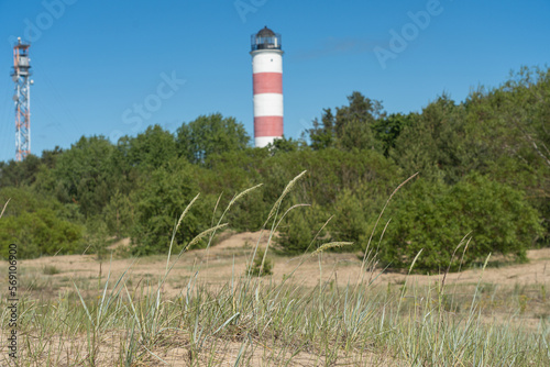 Narva-J  esuu  Estonia - 06.23.2022  Lighthouse on the coast. Green trees and a sandy beach overgrown with grass in the foreground. A sunny day.