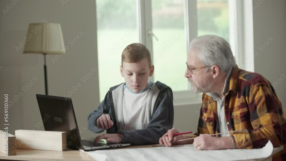 Grandpa is planning repairs and making notes in project with penci. Grandson is helping granddad solve complex calculation using laptop. Elderly man and young guy are sitting against background of