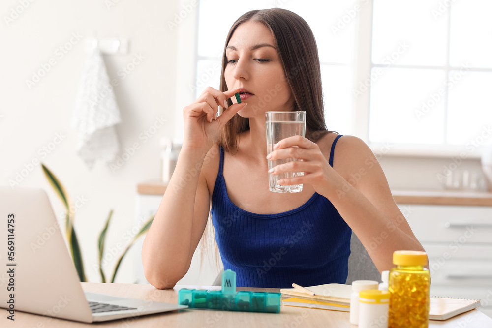 Beautiful woman with glass of water taking vitamin supplement in kitchen