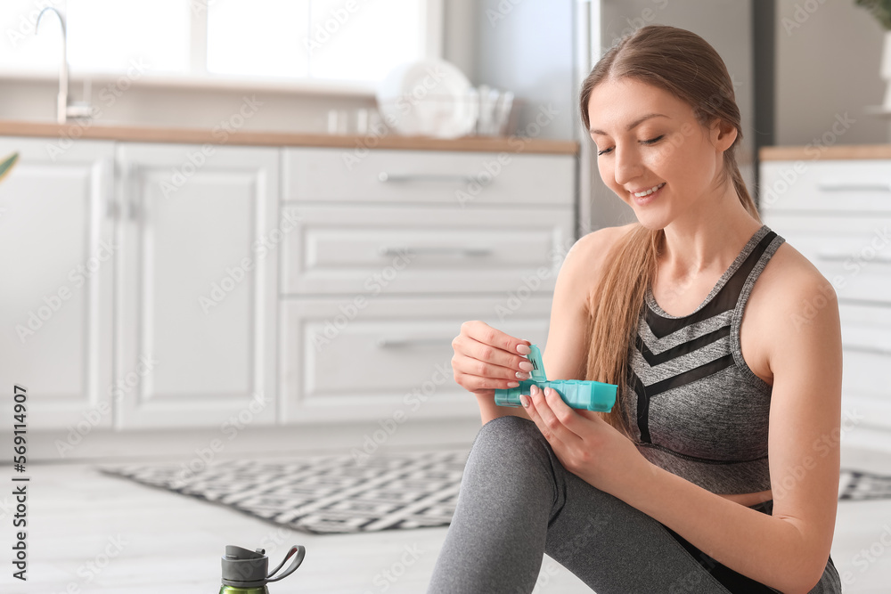 Sporty young woman with container of vitamin supplements in kitchen Sporty young woman with container of vitamin supplements in kitchen