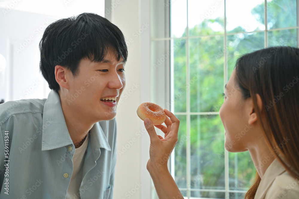 Young woman feeding his boyfriends with donut while cooking in modern kitchen at home, enjoying leisure weekend time together Young woman feeding his boyfriends with donut while cooking in modern kitchen at home, enjoying leisure weekend time together