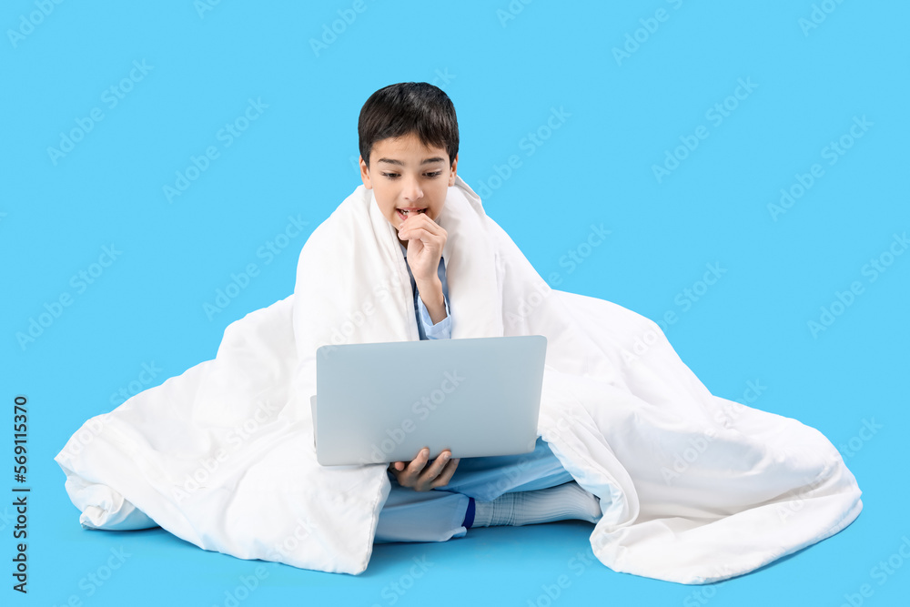 Little boy with blanket and laptop biting nails on blue background Little boy with blanket and laptop biting nails on blue background